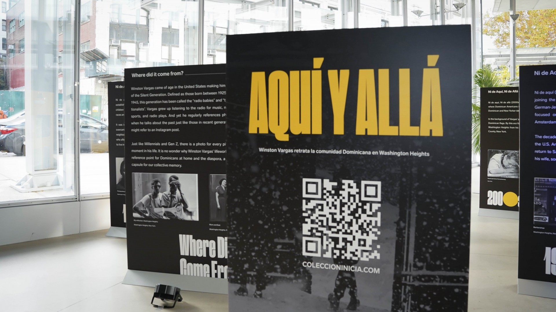 Large black foam-core boards displayed in The Forum's windowed Atrium as part of the Aqui y Alla exhibit. The boards feature photos and text.