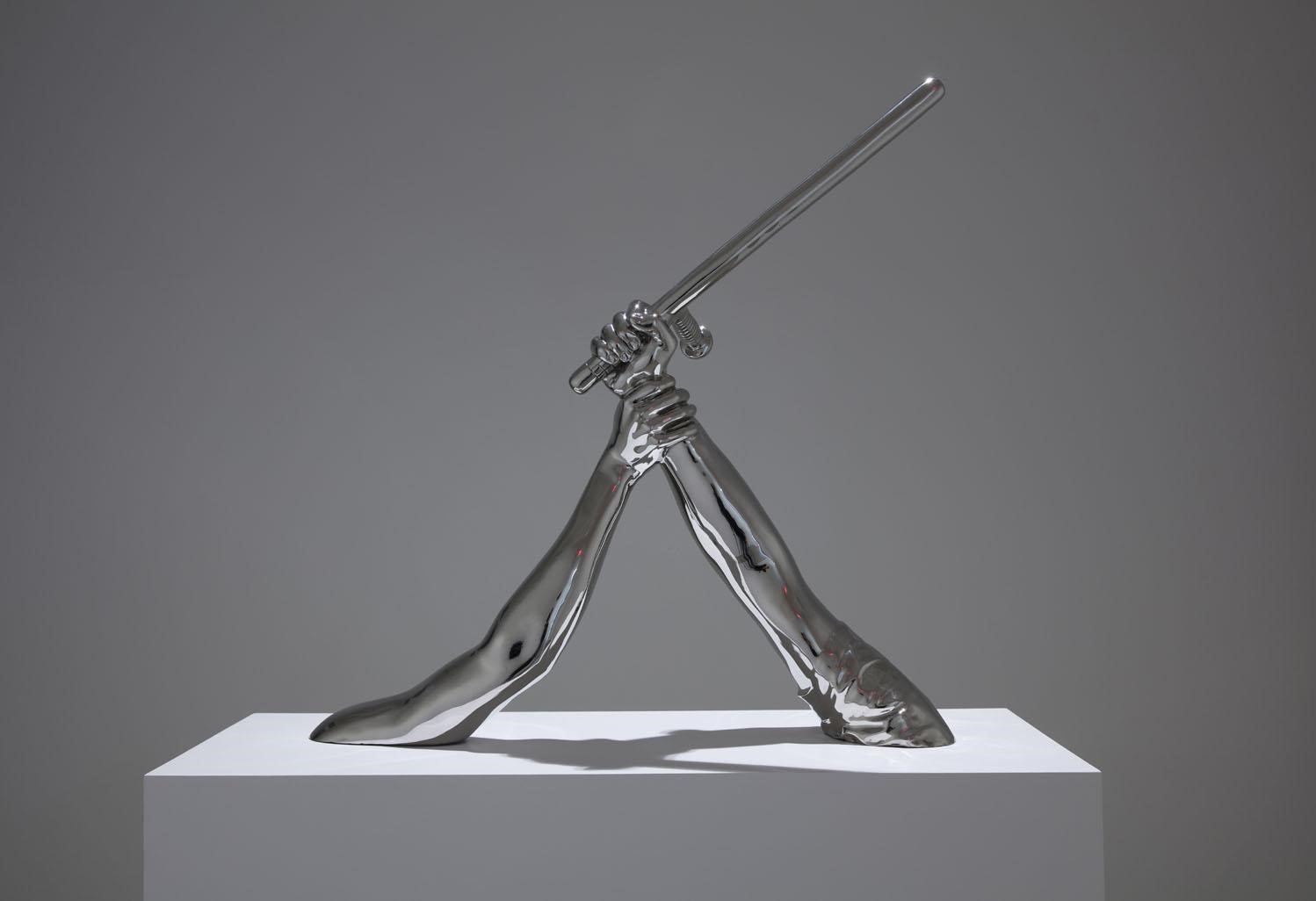 Photo of a metal sculpture by Hank Willis Thomas entitled Strike. It depicts two arms protuding from a white base. One arm is out-stretched and holding a baton-like instrument. The second arm is also reaching out, and has grasped the wrist of the other arm. 