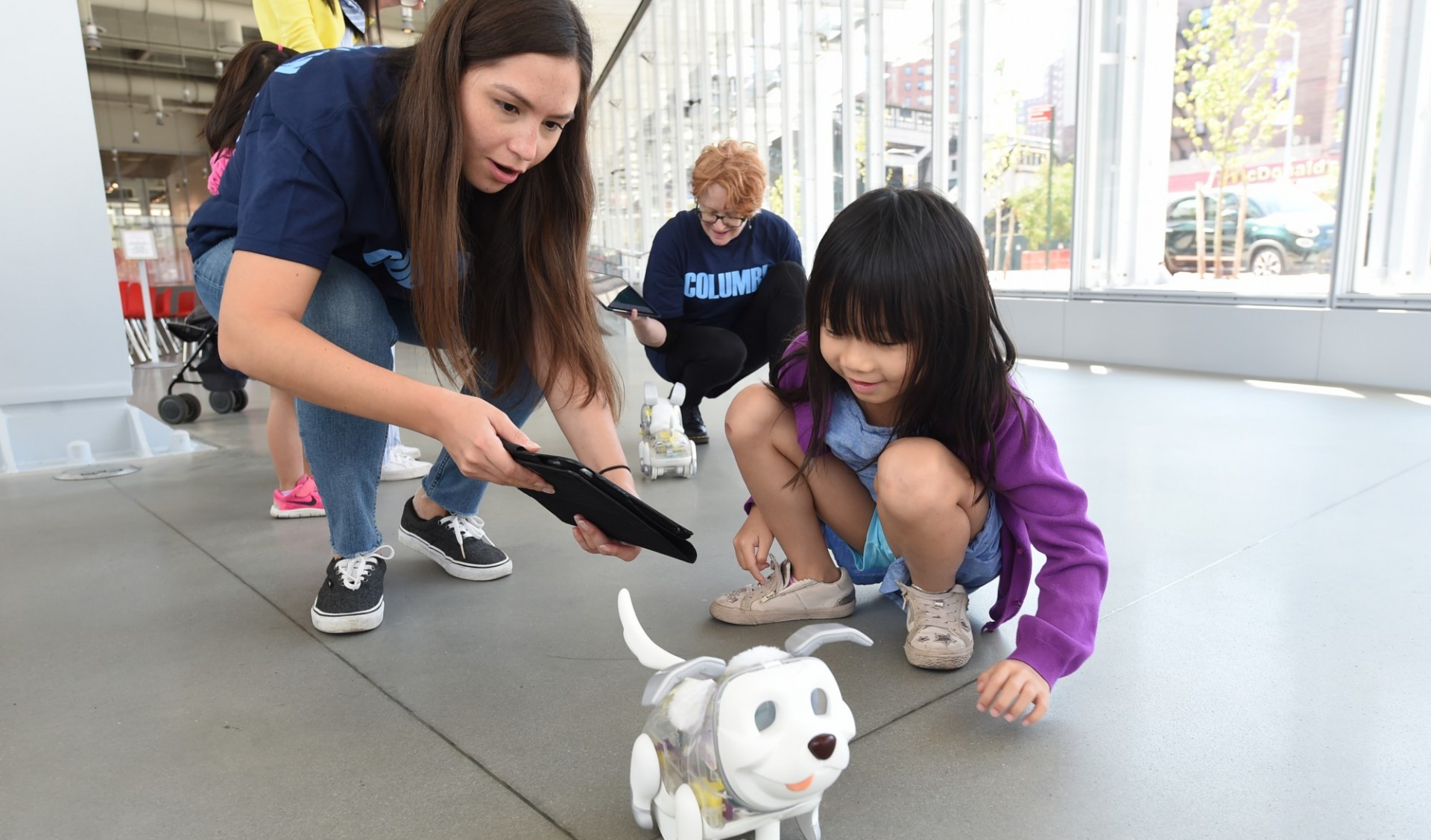 Young woman demonstrates how to control a robotic dog using an iPad while a young child looks on