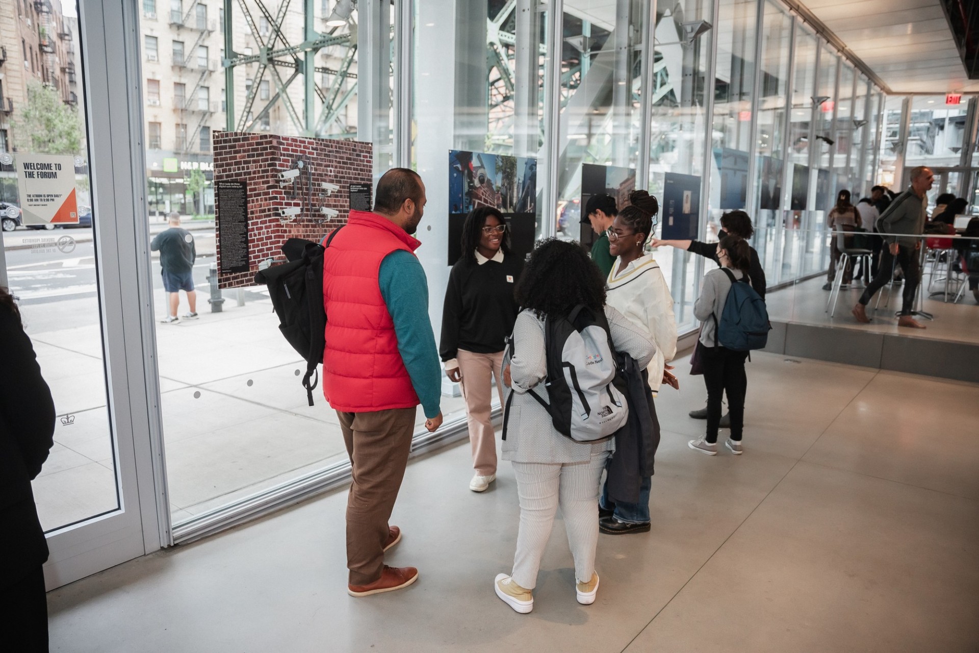 Group of students in conversation in front of the photo exhibit they helped create. Photo boards are hung in the building's windows as visitors look on.