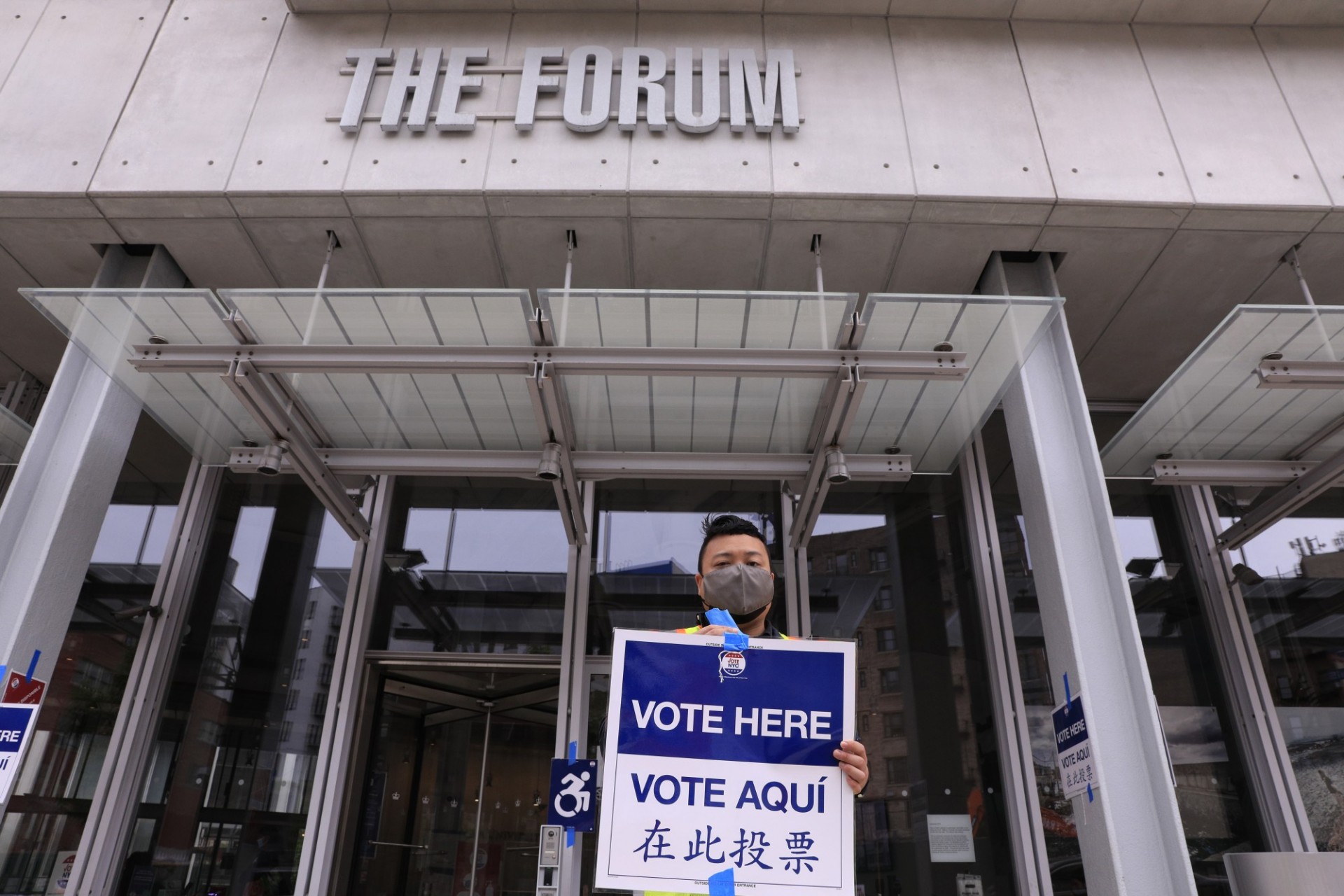 A man wearing a face mask stands in front of The Forum building holding a blue and white sign that says "Vote Here, Vote Aqui"