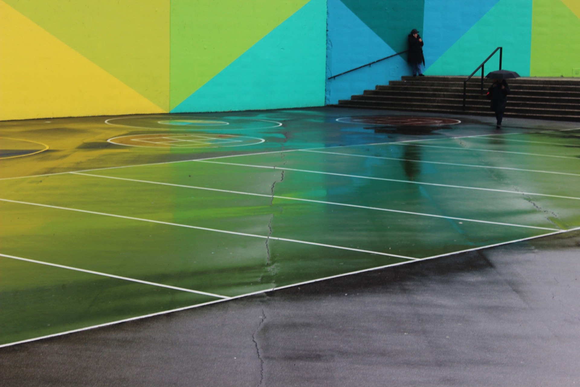 Image of a large open space with colorful, geometric patterns on the walls and floor. One person leans against the wall along a staircase. Another approaches the stairs holding an umbrella.