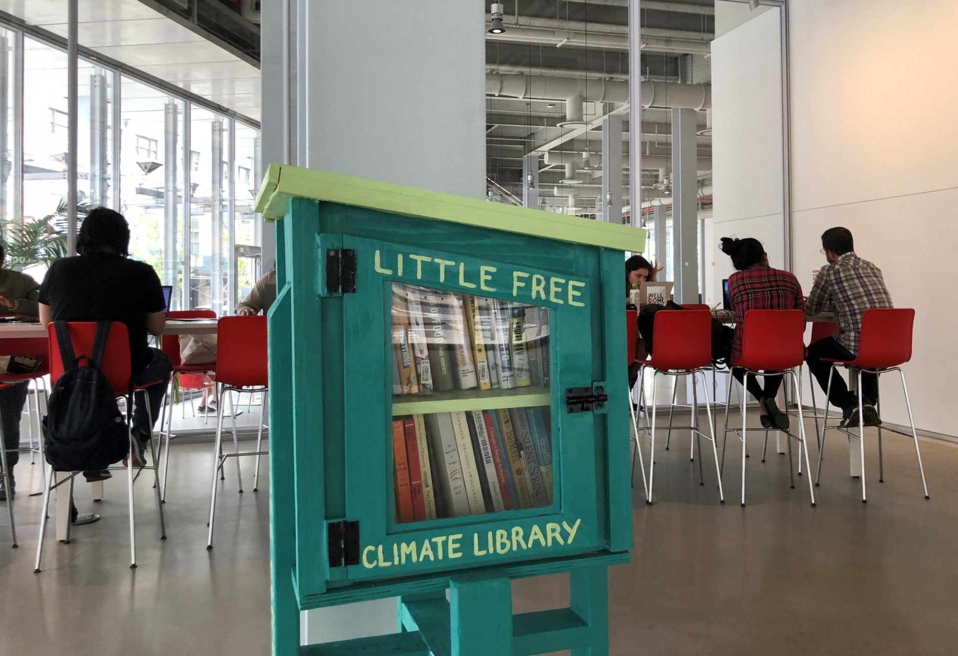 image of the Little Free Climate Justice Library in the Forum's Atrium, with people seated, studying in the background. The Library is a green wooden cupboard with a clear glass door, and inside, two shelves full of books. 