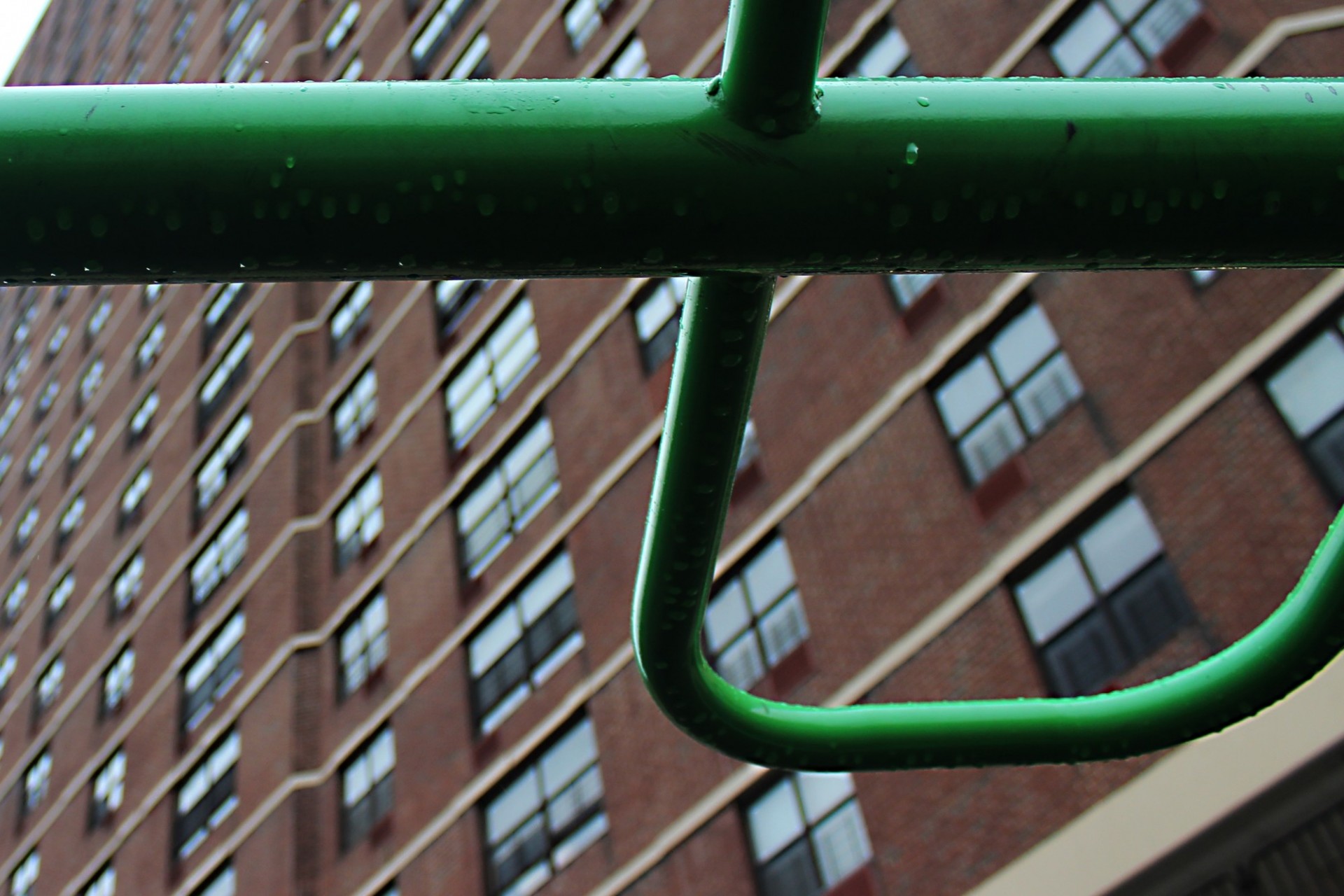 Close-up of raindrops on a green metal cross-bar, perhaps on a playground. A large apartment building with many windows looms in the background. 