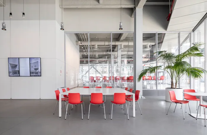 Large white square table in the Forum's atrium. Each side of the table has three red chairs. To the right of the table is another smaller table and a large plant. To the left, is a three-paneled digital screen. 