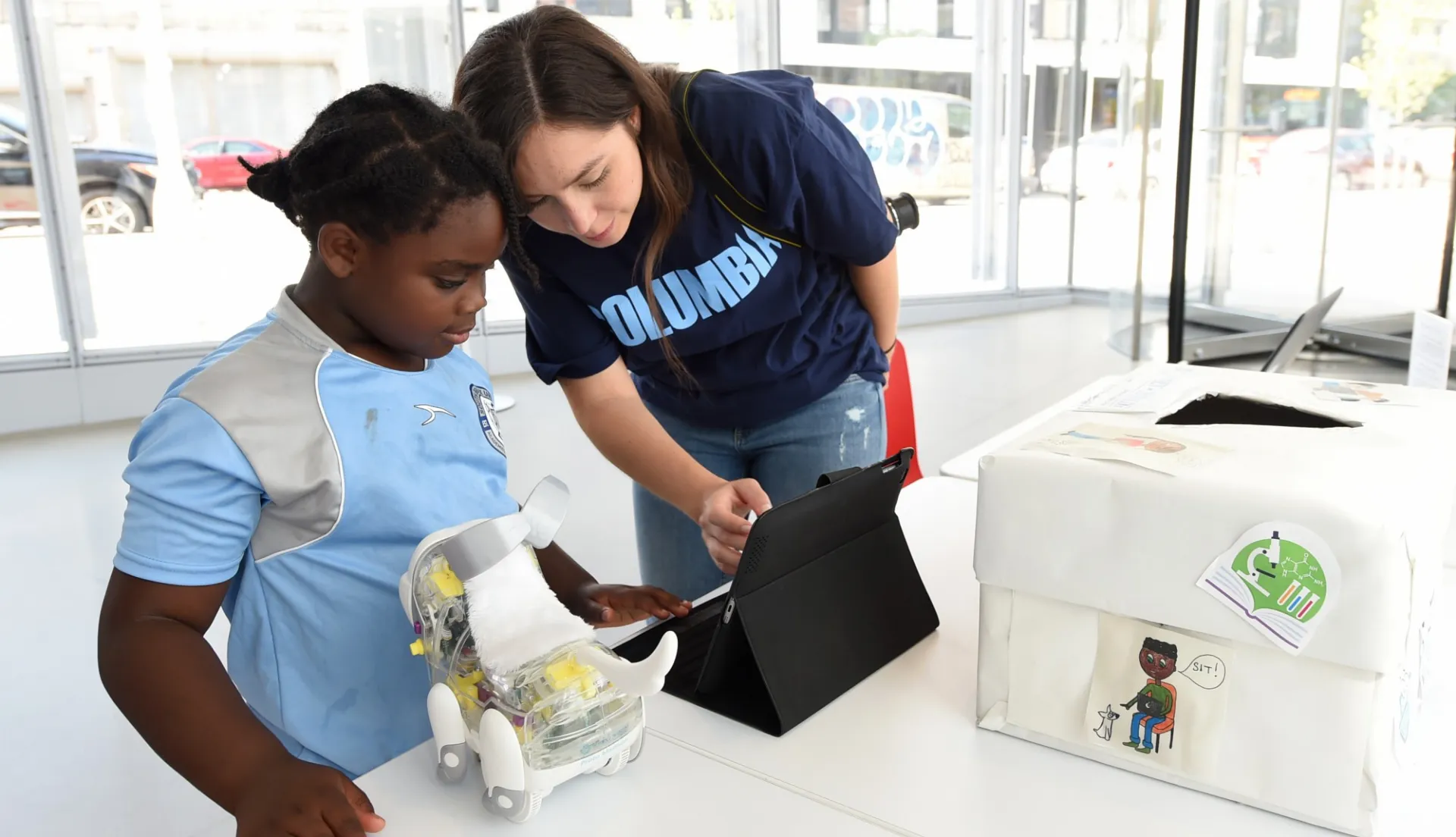 Young woman demonstrates how to control a robotic dog using an iPad while a young child looks on