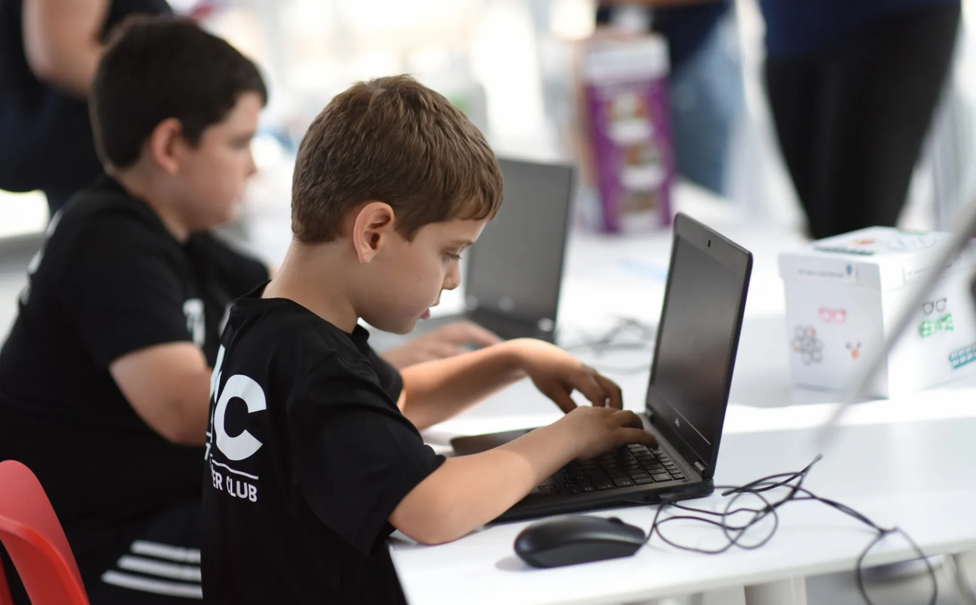 Two young boys sit side by side typing on laptops