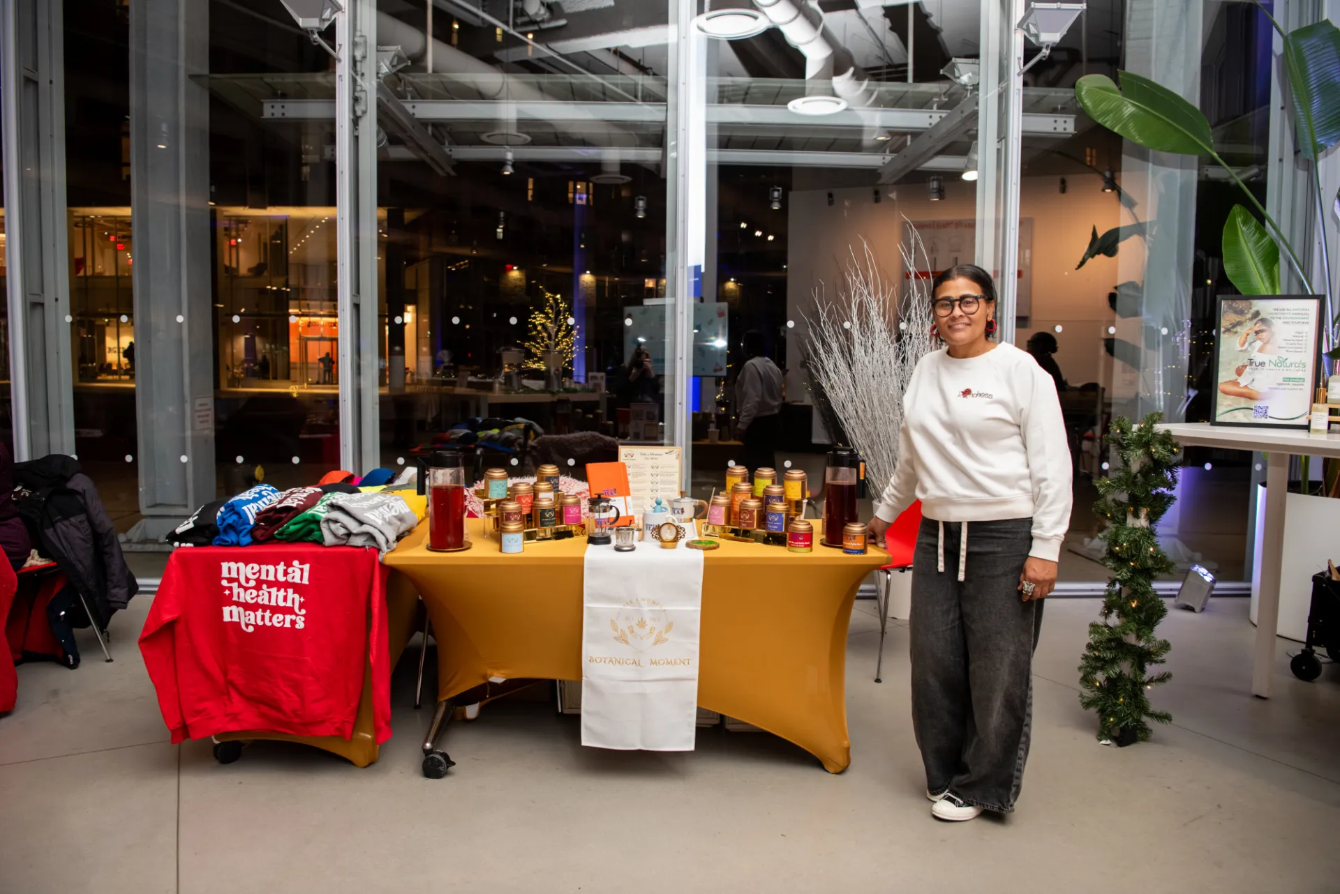 A holiday market vendor poses next to a booth featuring candles, teas, apparel, and wellness products inside the Lee C. Bollinger Forum. Floor-to-ceiling windows, winter décor, and warm lighting create a welcoming seasonal atmosphere.