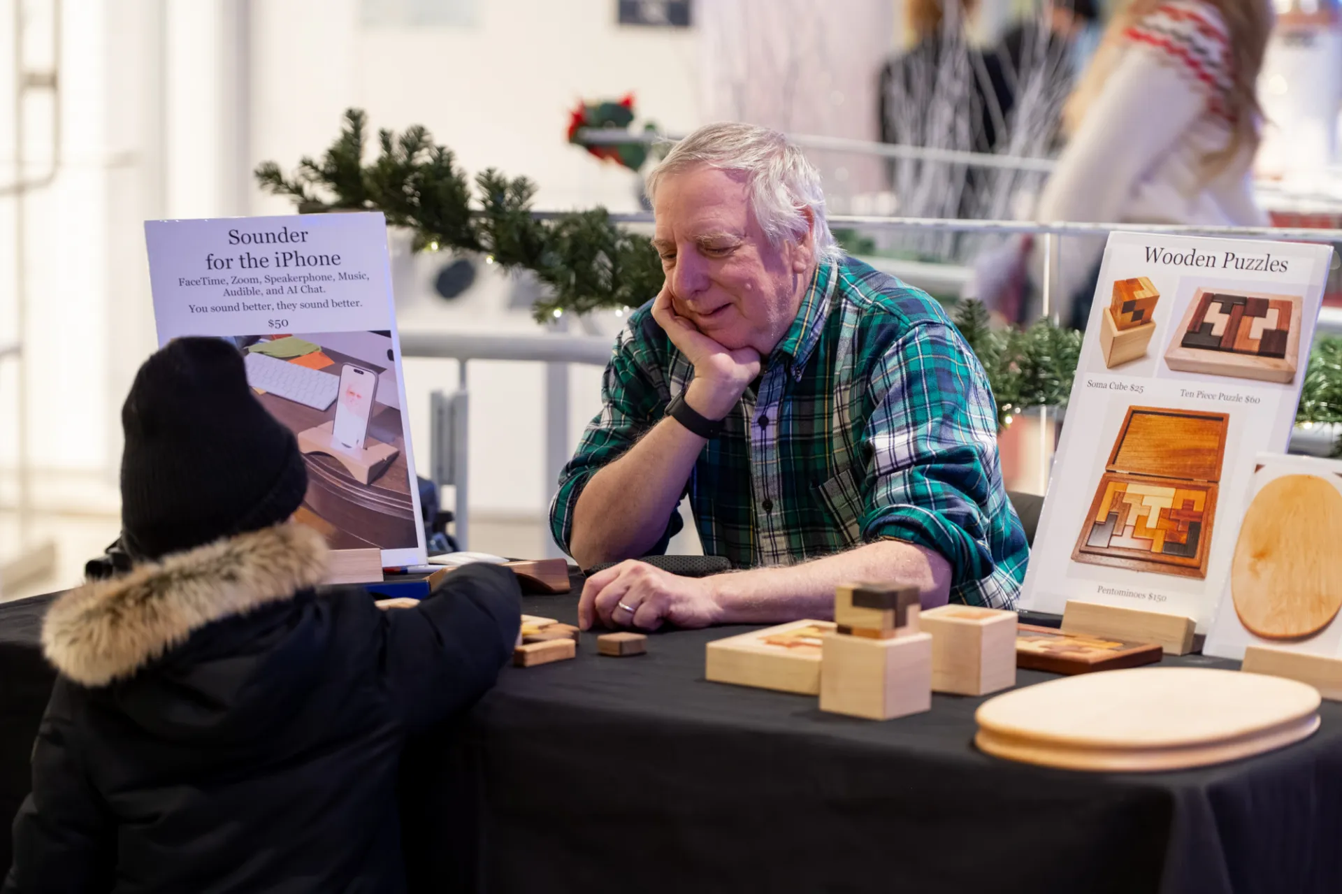 At an indoor holiday market, a vendor in a plaid shirt sits behind a display table covered with wooden puzzles, blocks, and circular wooden boards. A child wearing a dark winter coat with a fur-trimmed hood stands at the table looking at the items. Signs on the table advertise wooden puzzles and a smartphone sound amplifier, while holiday garland with lights decorates the booth in the background.