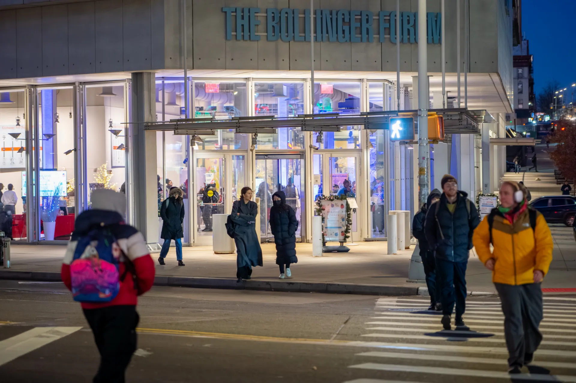 Exterior view of the Lee C. Bollinger Forum at dusk during a holiday market, with pedestrians crossing nearby and guests gathering at the brightly lit entrance. Holiday decorations and vendor tables can be seen through the building’s glass façade.