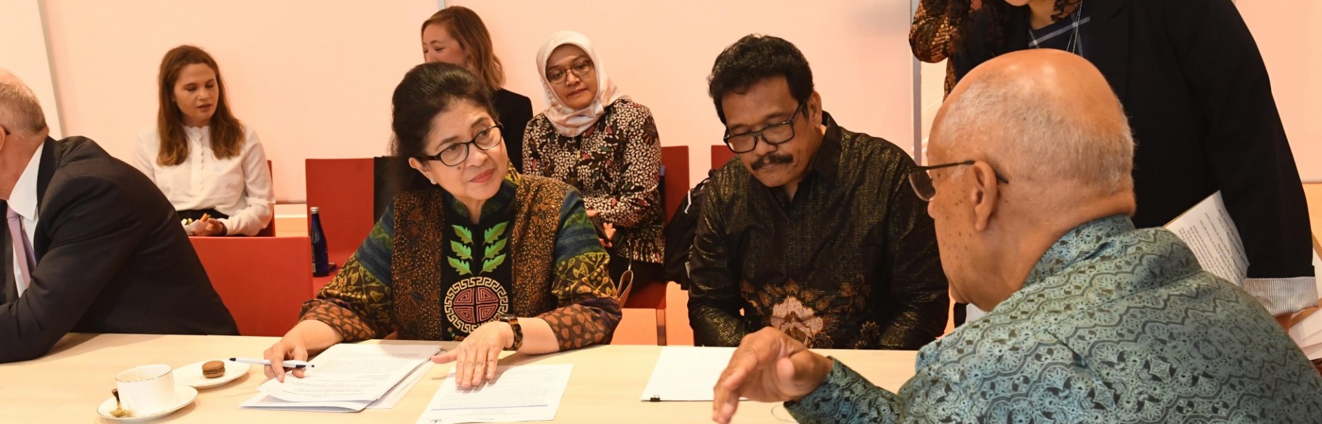 Three people sit around a conference table in discussion with one another. A woman stands above them, and three people sit in the background observing the conversation.