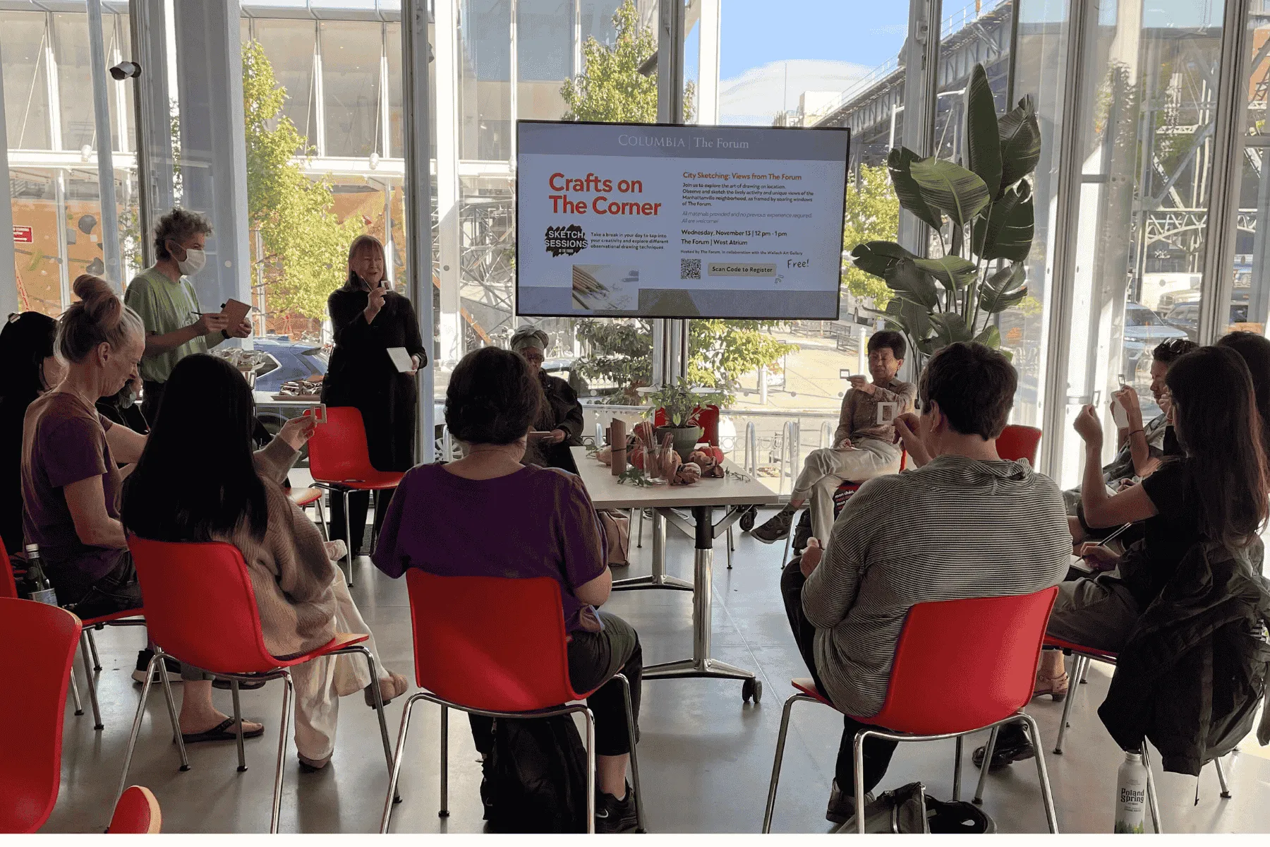 People sit in a circle on red chairs at Columbia’s Forum for a Crafts on the Corner sketching event. An educator stands near a large screen displaying event details.