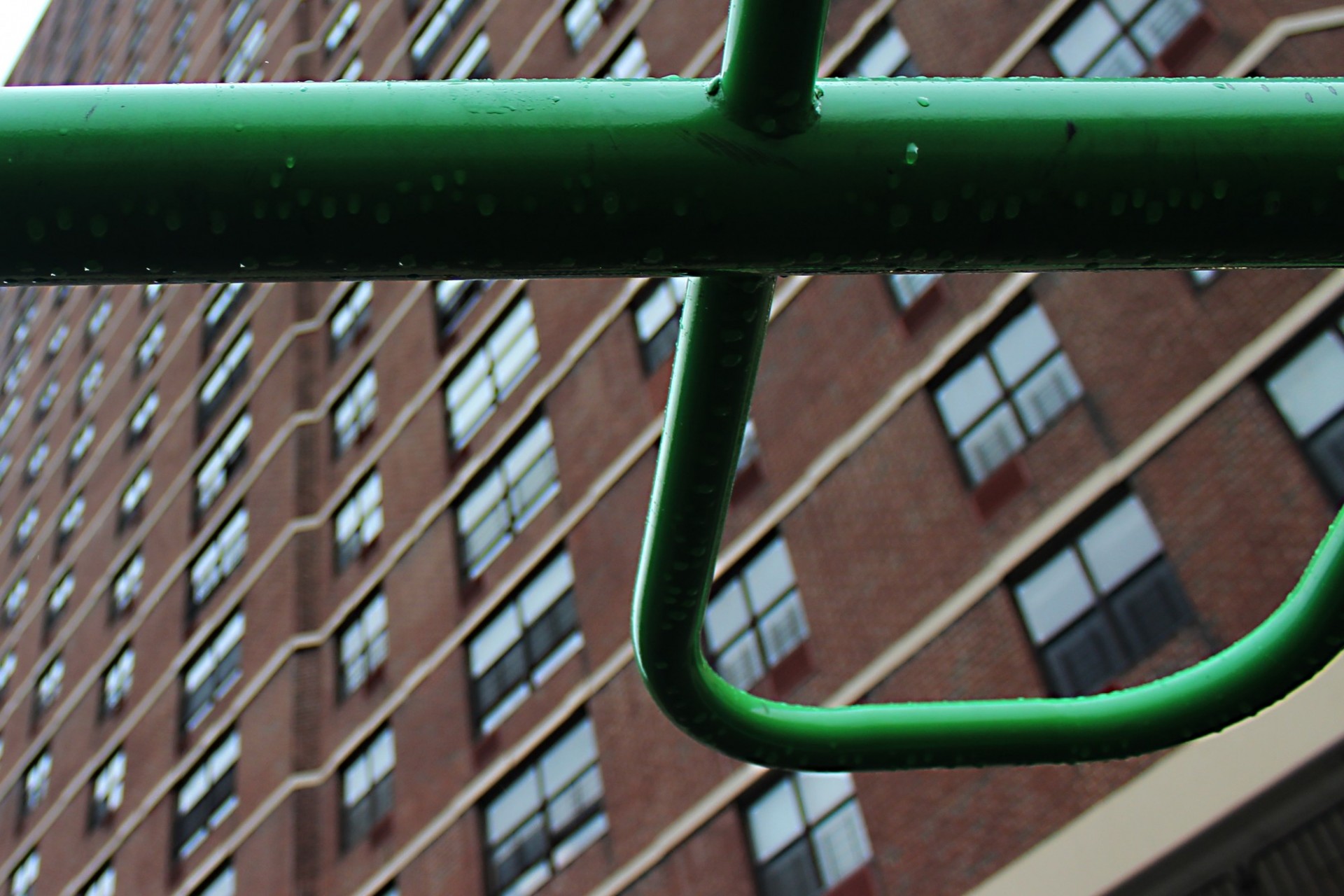 Photograph by Tyler Close-up of raindrops on a green metal cross-bar, perhaps on a playground. A large apartment building with many windows looms in the background.